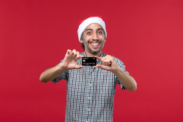 Vue de face jeune homme tenant une carte bancaire et souriant sur fond rouge