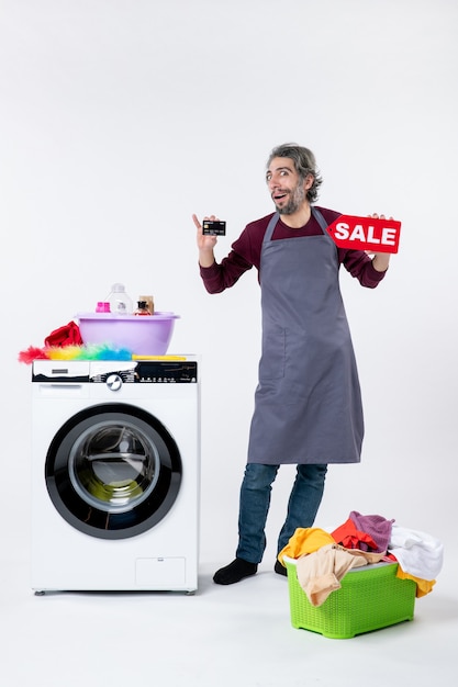 Photo gratuite vue de face jeune homme tenant une carte et un signe de vente debout près de la machine à laver sur un mur blanc