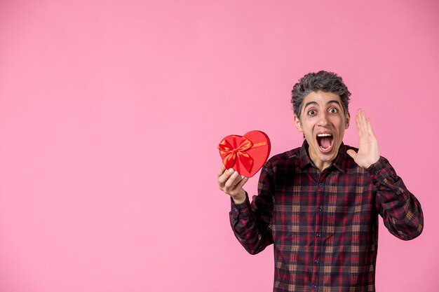 Vue de face jeune homme tenant un coeur rouge présent sur un mur rose clair