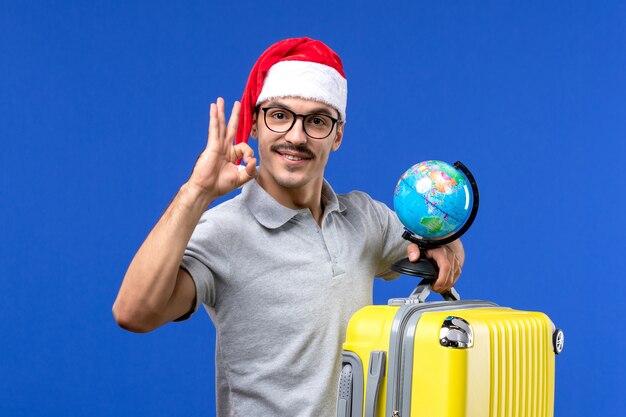 Vue de face jeune homme tenant globe et sac jaune sur voyage de vacances avion mur bleu
