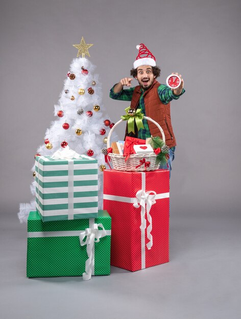 Vue de face jeune homme tenant une horloge autour de cadeaux sur sol gris cadeau nouvel an noël