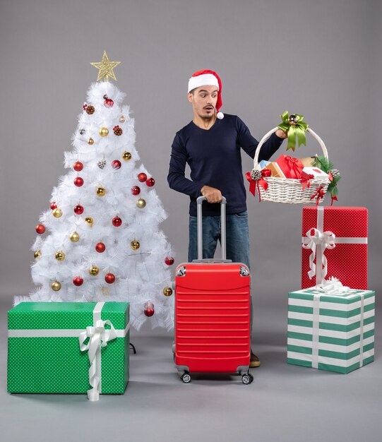 Vue de face jeune homme tenant un panier-cadeau et une valise debout près d'un arbre de Noël blanc et des cadeaux colorés sur gris isolé