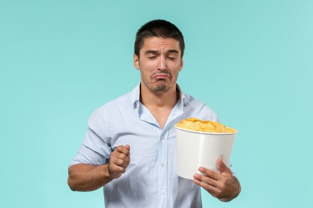 Vue de face jeune homme tenant un panier avec des cips de pommes de terre et regarder un film sur un mur bleu clair cinéma films à distance