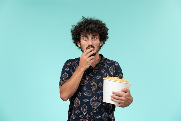 Vue de face jeune homme tenant un panier avec des cips de pommes de terre et regarder un film sur le mur bleu film cinéma masculin cinéma cinéma