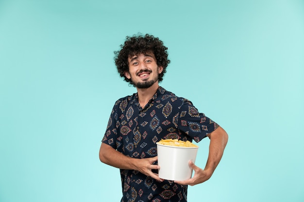 Vue de face jeune homme tenant un panier avec des cips de pommes de terre et souriant sur un mur bleu cinéma cinéma à distance cinéma cinéma