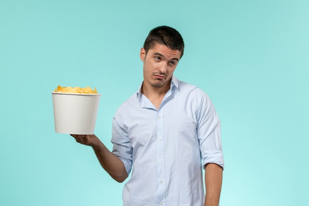 Vue de face jeune homme tenant le panier avec des croustilles sur une surface bleu clair