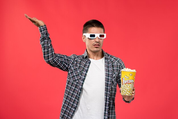 Vue de face jeune homme tenant un paquet de pop-corn et portant des lunettes de soleil -d sur un mur rouge clair cinéma cinéma film cinéma