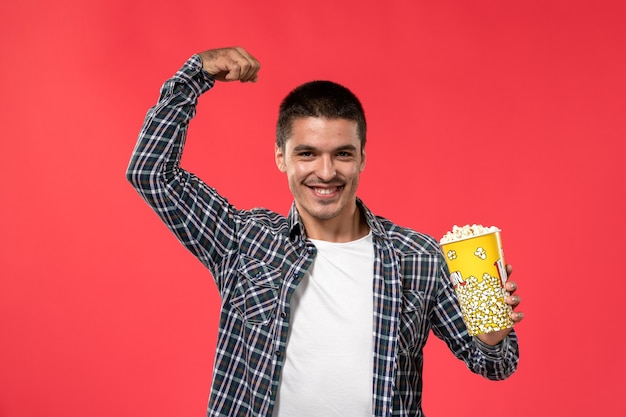 Vue de face jeune homme tenant le paquet de pop-corn et souriant flexion sur mur rouge clair cinéma films cinéma cinéma garçon garçon