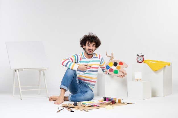 Vue de face d'un jeune homme tenant des peintures et un gland pour dessiner sur un mur blanc