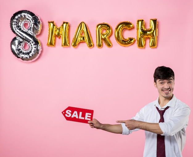 Vue de face jeune homme tenant une plaque signalétique de vente rouge avec décoration de mars sur fond rose fête argent féminin shopping homme présent vacances égalité des couleurs