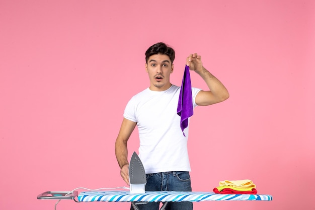 Vue de face jeune homme tenant une serviette et un fer à repasser sur fond rose effort de nettoyage vêtements émotion maison travaux ménagers homme travail