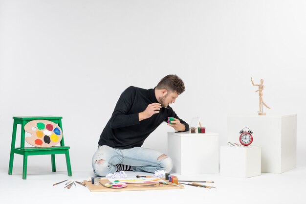 Vue de face jeune homme travaillant avec des peintures à l'intérieur de petites boîtes sur mur blanc artiste peinture art couleur peinture photo tirage photo