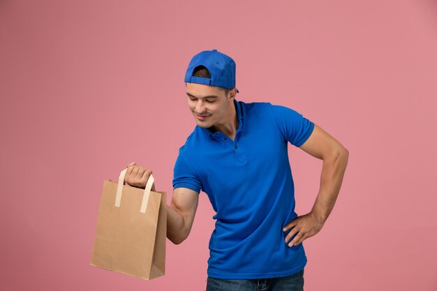 Vue de face jeune homme en uniforme bleu et cape avec paquet de papier de livraison sur ses mains sur le mur rose