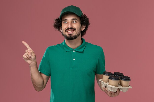 Photo gratuite vue de face jeune homme en uniforme vert et cape tenant des tasses de café de livraison marron sur le fond rose service travail de livraison uniforme homme