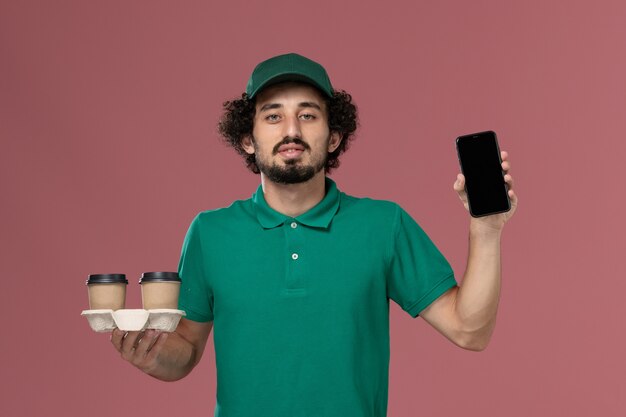 Vue de face jeune homme en uniforme vert et cape tenant des tasses de café de livraison et son téléphone sur le travail de service de fond rose travailleur de la livraison uniforme
