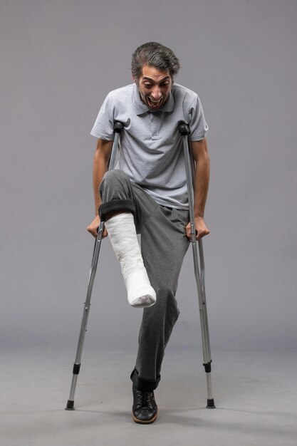 Vue de face jeune homme utilisant des béquilles en raison d'un pied cassé sur le mur gris désactiver l'accident cassé du pied de jambe