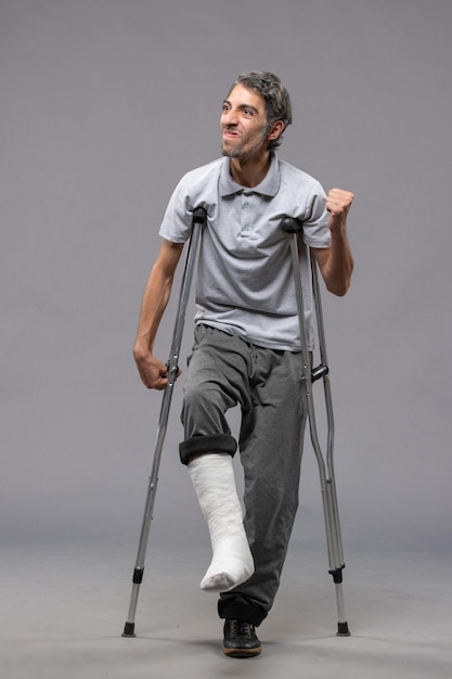 Vue de face jeune homme utilisant des béquilles en raison d'un pied cassé sur un mur gris pied handicapé jambe cassée accident