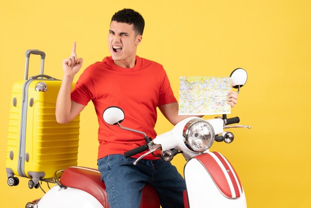 Vue de face jeune homme en vêtements décontractés sur cyclomoteur tenant la carte de voyage pointant le doigt vers le haut