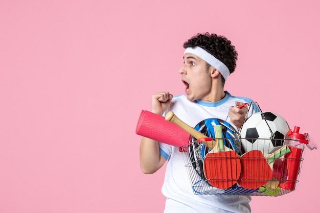 Vue de face jeune homme en vêtements de sport avec panier plein de choses de sport mur rose