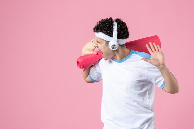 Vue de face jeune homme en vêtements de sport avec tapis de yoga et écouteurs sur mur rose