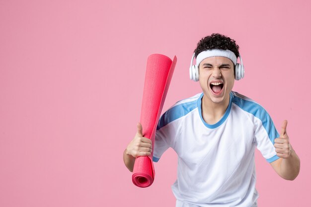 Vue de face jeune homme en vêtements de sport avec tapis de yoga sur mur rose