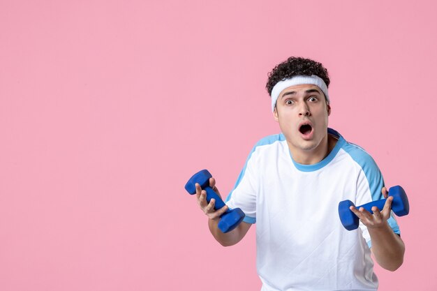 Vue de face jeune homme en vêtements de sport travaillant avec des haltères sur un mur rose