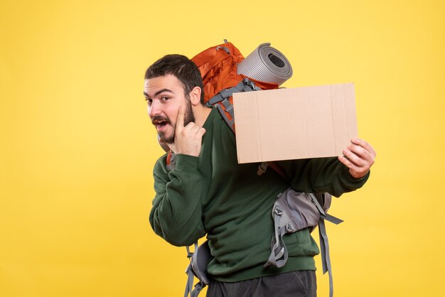 Vue de face d'un jeune homme de voyage confiant avec sac à dos et montrant une feuille sans écrire sur fond jaune