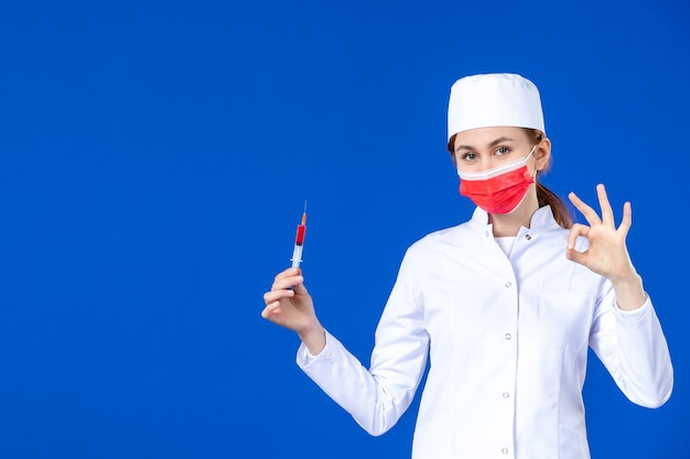 Vue de face jeune infirmière en costume médical blanc avec masque rouge et injection dans ses mains sur le mur bleu