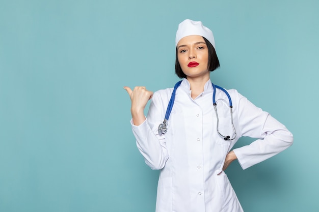 Une vue de face jeune infirmière en costume médical blanc et stéthoscope bleu posant sur le bureau bleu couleur médecin hôpital médecine