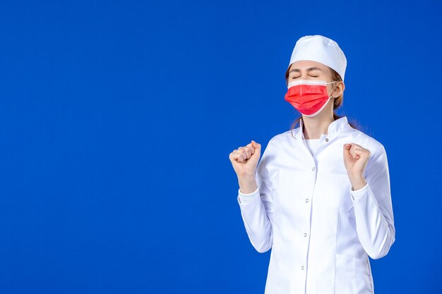 Vue de face jeune infirmière en costume médical avec masque de protection rouge sur mur bleu