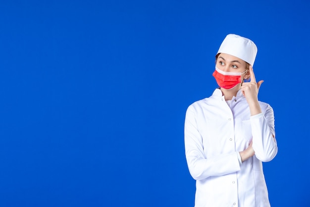 Vue de face jeune infirmière en costume médical avec masque de protection rouge sur mur bleu