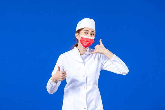 Vue de face jeune infirmière en costume médical avec masque de protection rouge sur mur bleu