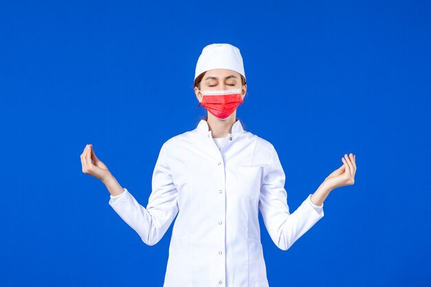 Vue de face jeune infirmière en costume médical avec masque de protection rouge sur mur bleu