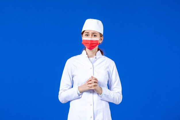 Vue de face jeune infirmière en costume médical avec masque de protection rouge sur mur bleu