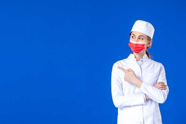 Vue de face jeune infirmière en costume médical avec masque de protection rouge sur mur bleu