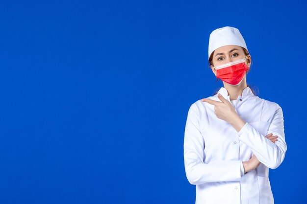 Vue de face jeune infirmière en costume médical avec masque de protection rouge sur mur bleu