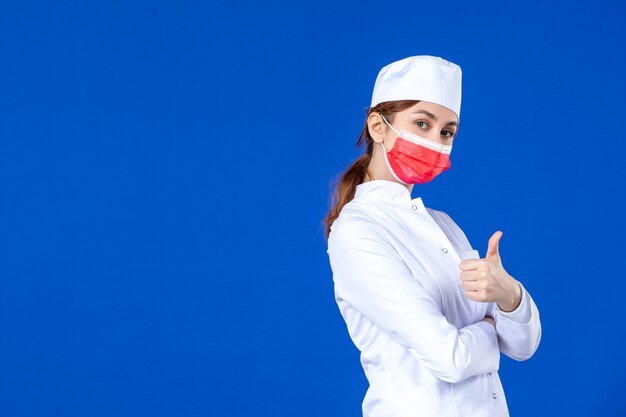Vue de face jeune infirmière en costume médical avec masque rouge sur mur bleu