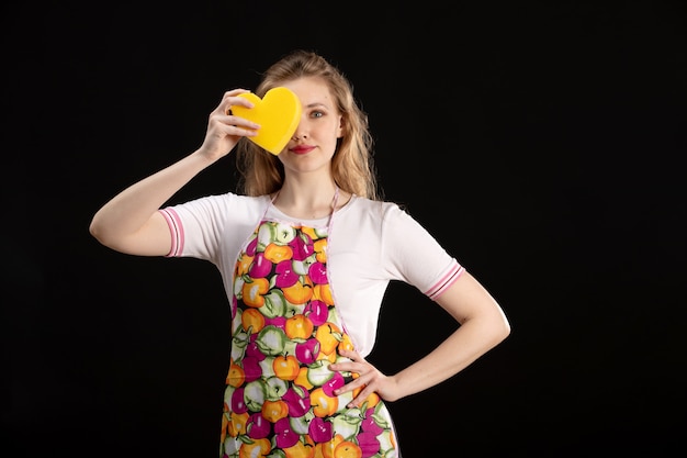 Photo gratuite une vue de face jeune jolie fille en cape colorée souriant tenant en forme de cœur jaune sur le fond noir amour sourire positivité