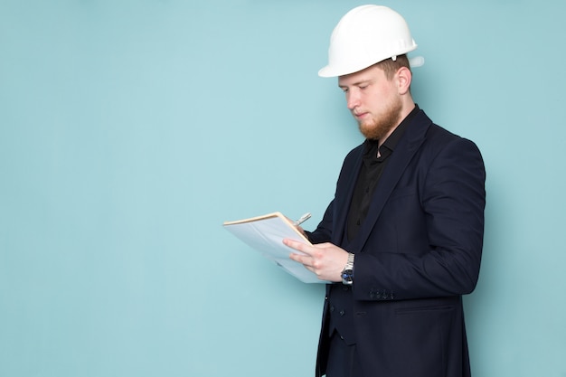 Une vue de face jeune mâle attrayant avec barbe en costume moderne classique noir foncé en casque de construction blanc écrire des notes sur l'espace bleu