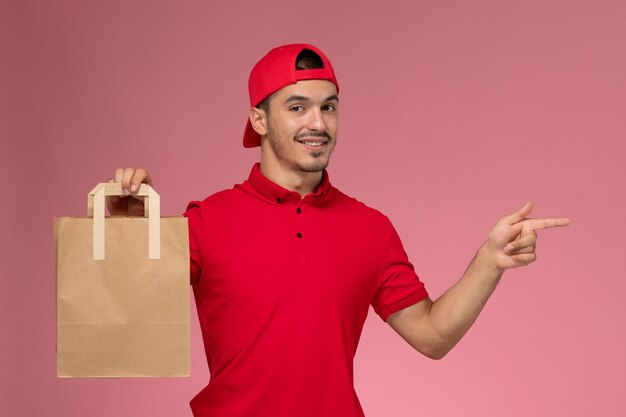 Vue de face jeune messager mâle en cape uniforme rouge tenant un paquet de papier alimentaire sur le fond rose clair.