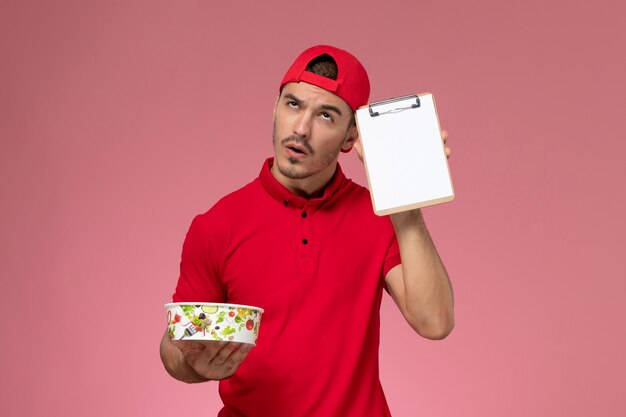 Vue de face jeune messager masculin en cape uniforme rouge tenant le bol de livraison rond et le bloc-notes pensant sur fond rose clair.