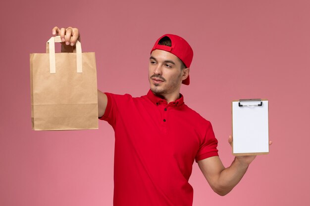 Vue de face jeune messager masculin en cape uniforme rouge tenant le paquet de nourriture et le bloc-notes sur fond rose.