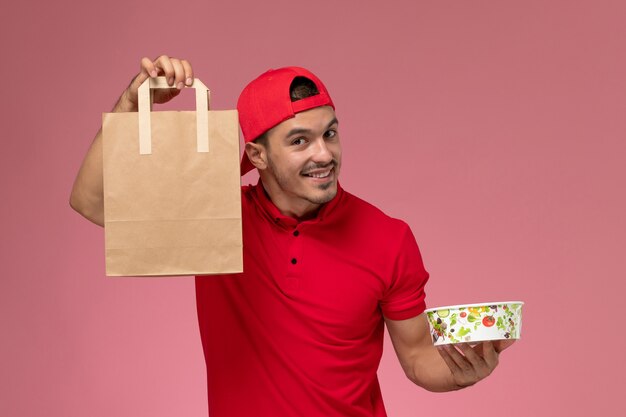 Vue de face jeune messager masculin en cape uniforme rouge tenant le paquet de nourriture et un bol avec le sourire sur le fond rose.