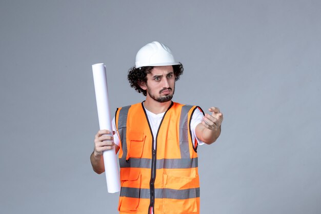 Vue de face d'un jeune ouvrier du bâtiment inquiet en gilet d'avertissement avec casque de sécurité et montrant un blanc sur un mur gris