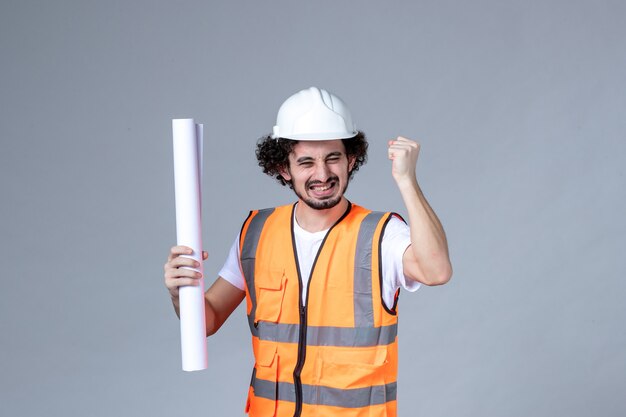 Vue de face d'un jeune ouvrier du bâtiment souriant et heureux en gilet d'avertissement avec casque de sécurité et montrant un blanc sur un mur gris