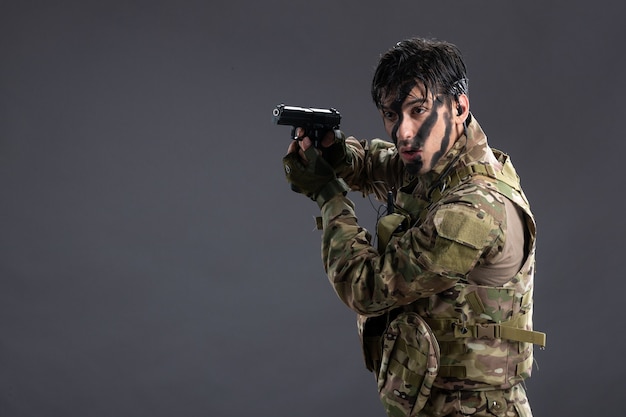 Vue de face jeune soldat combattant en tenue de camouflage avec arme à feu sur le mur sombre