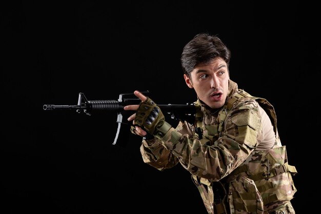 Vue de face jeune soldat en uniforme avec fusil sur le mur noir
