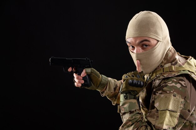 Vue de face jeune soldat en uniforme visant le pistolet sur mur noir