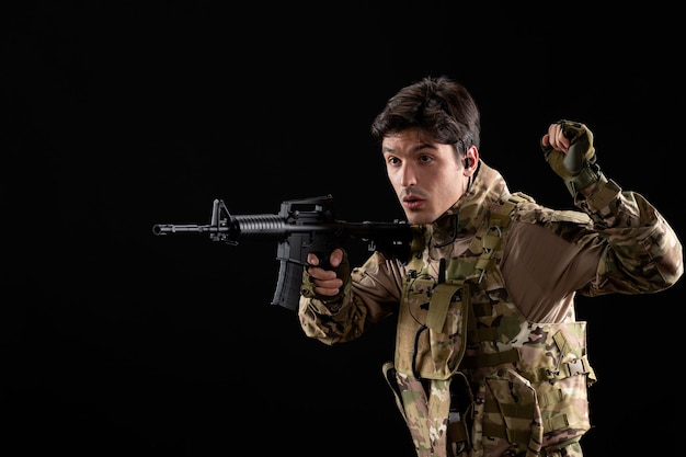 Vue de face jeune soldat en uniforme visant son fusil sur mur noir