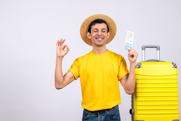 Vue de face jeune touriste debout près de valise jaune faisant signe ok tenant un billet